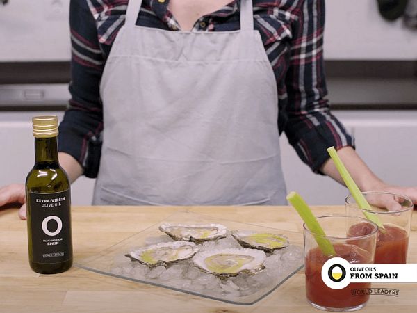 Wooden table with bottle of olive oil, plate with ice and oysters and transparent glass cup with Bloddy Mary with celery brunch