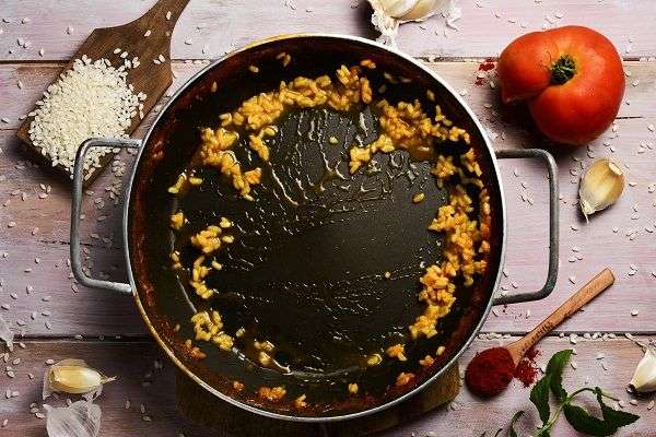 Paella pan with remains of yellow rice on wooden table with wooden spoon with raw rice, tomato and garlic cloves