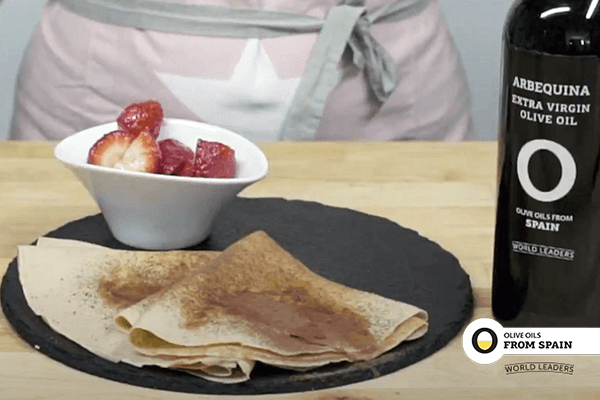 Slate plate with pancakes and a bowl of strawberries on wooden table. Next bottled olive oil from Spain. In the background person with apron.
