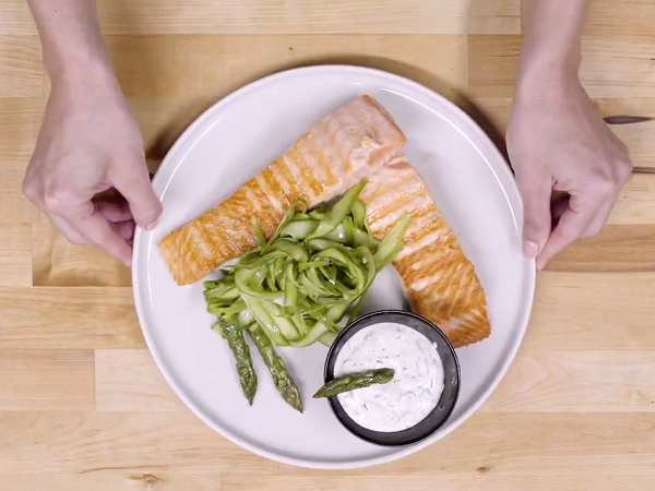 Hands showing white plate with grilled salmon, asparagus and yogurt sauce in a black bowl decorated with a green asparagus.