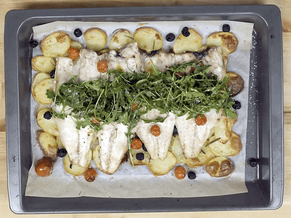 Baking tray on wooden table with sea bass with rosemary, black olives and cherry tomatoes on potato base