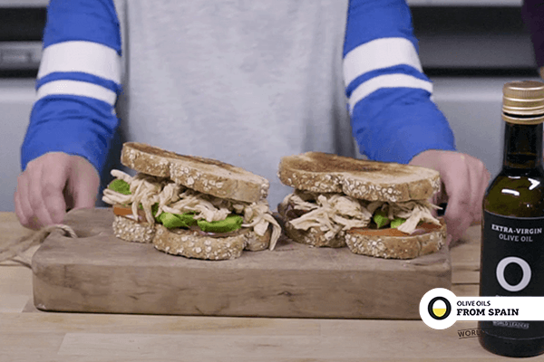 Child arms showing chicken sandwich with paprika and avocado on wooden board on wooden table with a bottle of olive oil